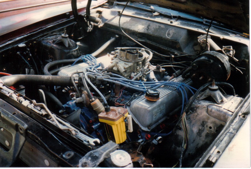 V8 engine bay with carburetor and blue spark plug wires installed in a car under the hood