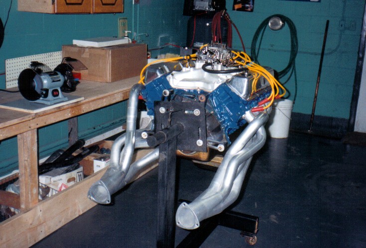 Blue V8 engine on a stand with chrome long-tube headers, bench grinder in a green-walled shop