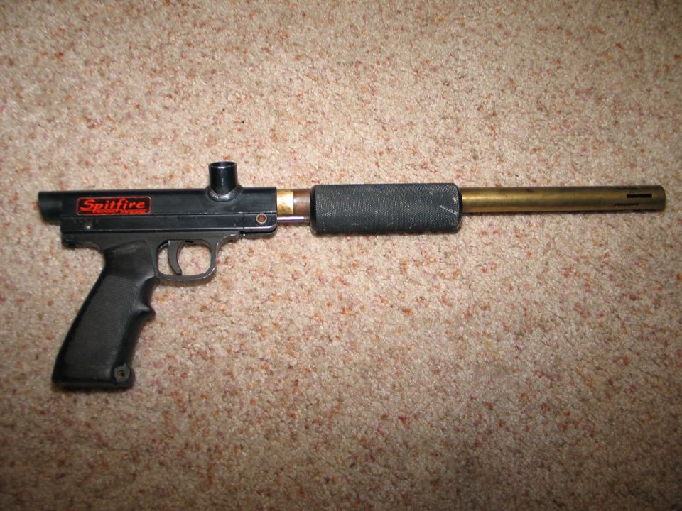 Black Spitfire paintball gun with brass barrel and textured grip, lying on carpet