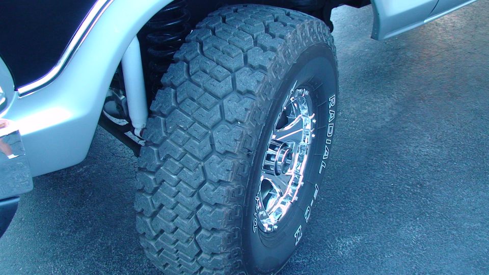 Close-up of a large off-road tire on a chrome wheel, with part of a silver vehicle body visible