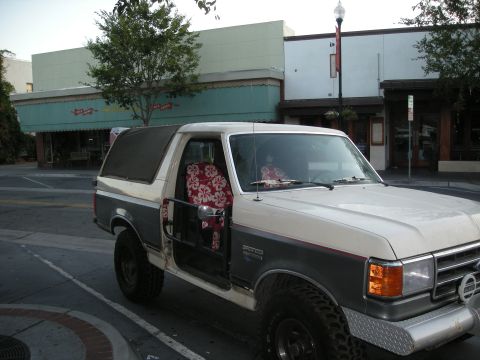 1991 Ford Bronco - A two-tone Ford Bronco is parked on a street in front of storefronts.
