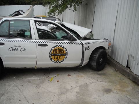 1996 Ford Crown Victoria - A damaged taxi cab sits parked next to a corrugated metal wall, showing significant damage to its front end.