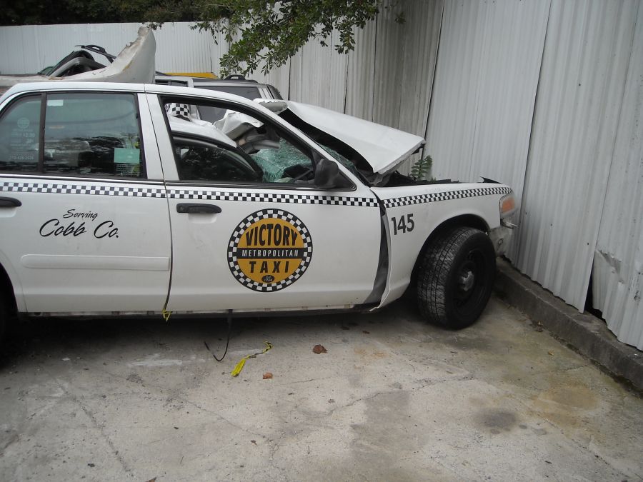White taxi cab with front end damage, hood crumpled, windshield cracked. "Victory Metropolitan Taxi" logo on door