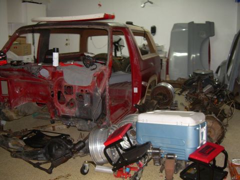 1986 Ford Bronco - A red SUV body is in a state of disassembly, with various automotive parts surrounding it in a garage setting.