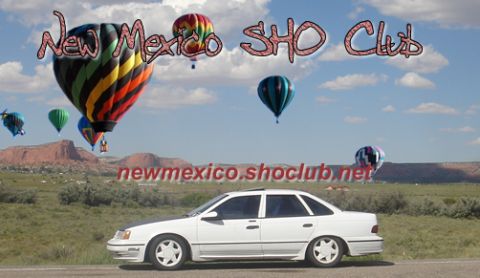 1990 Ford Taurus - A white Ford Taurus is parked on the side of a road with several hot air balloons in the sky above a desert landscape.