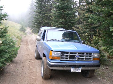 2006 Ford E350 - A blue pickup truck drives on a dirt road through a forest with fog in the distance.