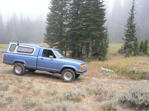 2006 Ford E350 - A blue pickup truck with a camper shell is parked on a dirt road surrounded by trees and fog.