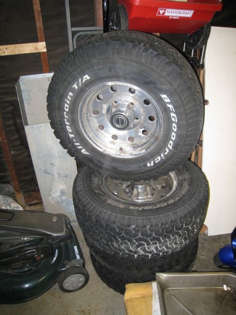 1996 Ford Bronco - Four BFGoodrich All-Terrain T/A tires stacked with rims. A red wheelbarrow is in the background.