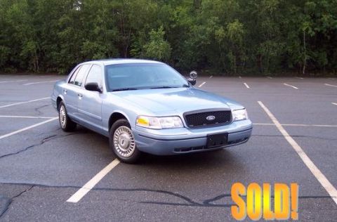 2001 Ford Crown Victoria - Light blue 2001 Ford Crown Victoria sedan parked in an empty lot with trees in the background.