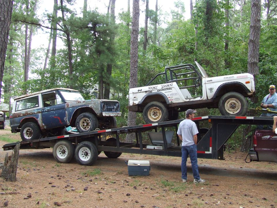Two rusty Ford Broncos on a trailer, one blue and one white, with men standing nearby