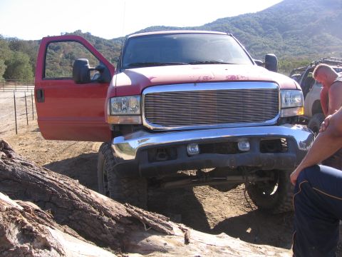 1989 Ford Bronco - A red Ford F-250 truck is parked off-road with its door open, showing a chrome grille and lifted suspension.