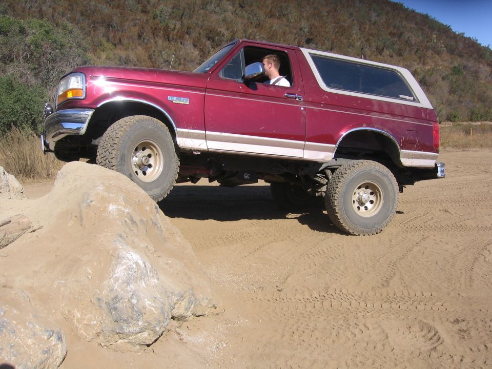 Red Ford Bronco with a white top driving over rocks on a dirt path
