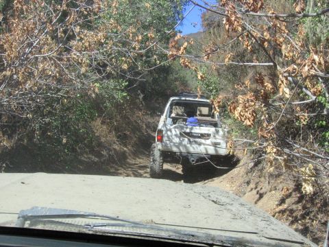 1989 Ford Bronco - A white Toyota pickup truck navigates a narrow dirt path surrounded by dry brush and trees.