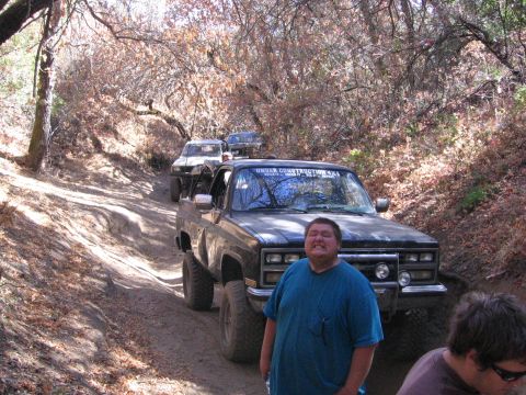 1989 Ford Bronco - Several off-road vehicles navigate a dusty, wooded trail on a sunny day.