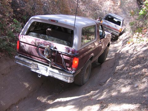 1989 Ford Bronco - Two SUVs navigate a narrow, rutted dirt path surrounded by trees.