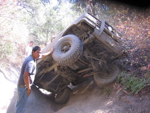 1989 Ford Bronco - A man attempts to right a heavily mud-covered off-road vehicle that has tipped onto its side on a dirt trail.