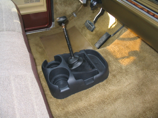 Interior view of a vehicle's floor console with a shifter and cup holders, showing pedals and part of a seat