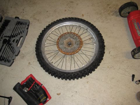 1993 Suzuki RMX250 - A motorcycle wheel and tire assembly is shown on a concrete floor with various tools and a lawnmower nearby.