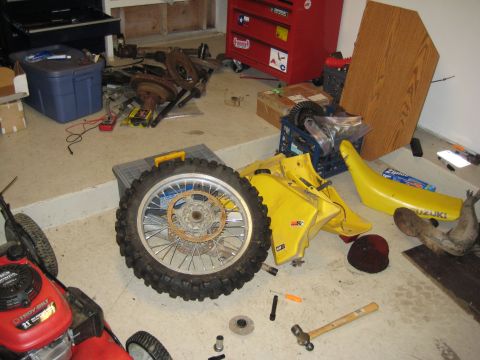 1993 Suzuki RMX250 - Disassembled yellow motorcycle parts, including a fuel tank, seat, and wheel, are laid out on a garage floor next to tools and a lawnmower.