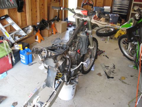 1993 Suzuki RMX250 - A motorcycle in a garage is partially disassembled, with its engine and suspension components visible. Another motorcycle is parked nearby.