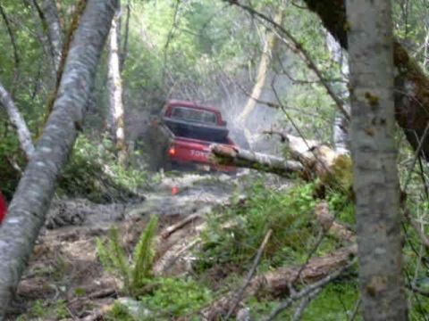 1988 Ford Bronco - A red Toyota pickup truck is seen off-roading on a challenging, muddy trail surrounded by dense forest.
