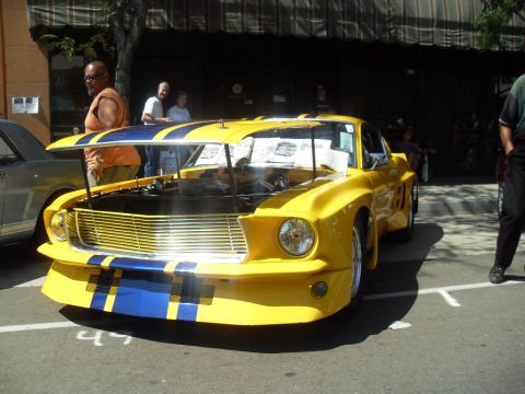 1990 Ford Bronco - A bright yellow 1967 Ford Mustang with blue racing stripes is displayed with its hood open, parked next to another vehicle.