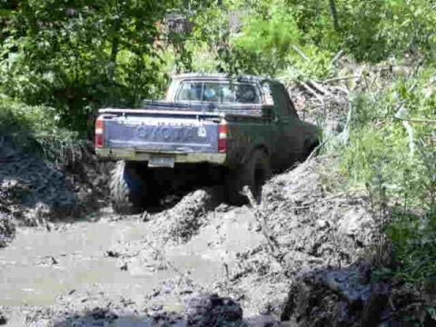 1988 Ford Bronco - A pickup truck navigates a muddy off-road trail, its rear end partially submerged in thick mud.