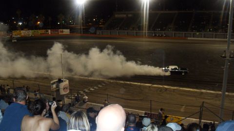 1990 Ford Bronco - A car performs a burnout on a race track at night, surrounded by spectators and illuminated by stadium lights.