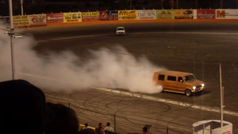 1990 Ford Bronco - An orange van drifts on a track, generating a large cloud of smoke, with another van visible in the distance.