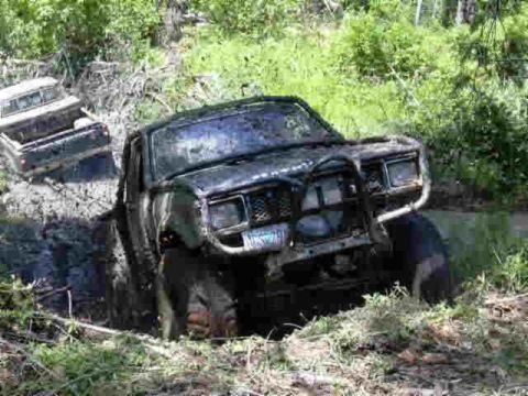 1988 Ford Bronco - Two trucks are stuck in a muddy, wooded area. The front vehicle is partially submerged in mud.