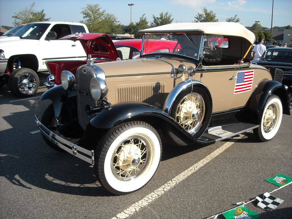 Tan convertible with white top and an American flag decal on the side. Other vehicles are parked nearby