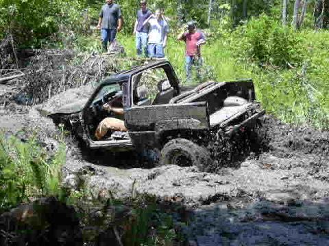 1988 Ford Bronco - A black SUV is deeply stuck in a muddy off-road trail, with several onlookers observing the scene.