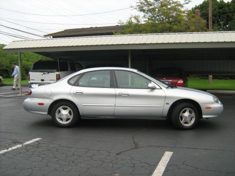 1988 Mercury Grand Marquis - A silver sedan is parked under a carport next to a white SUV. A person in a light blue shirt walks away from the vehicles.