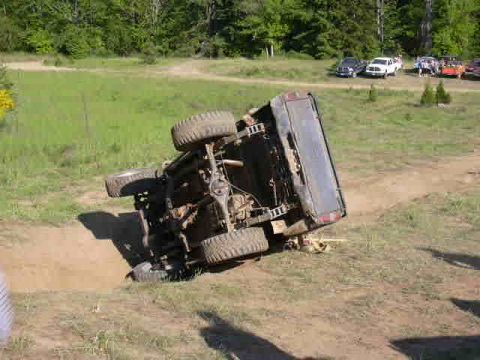 1988 Ford Bronco - An overturned SUV rests on its side in a dirt area, with several other vehicles parked nearby.