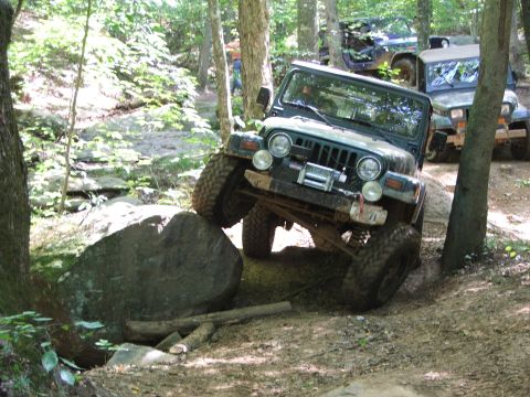 1996 Ford Bronco - A Jeep Wrangler maneuvers over a large rock on a wooded trail, with other off-road vehicles visible in the background.