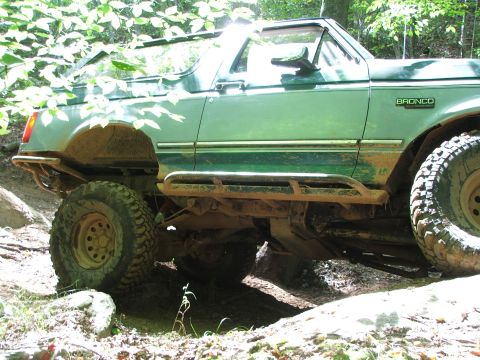 1996 Ford Bronco - Green 1996 Ford Bronco navigating a muddy, wooded trail with aftermarket side steps and bumpers.