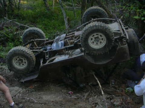 1988 Ford Bronco - An off-road vehicle has rolled onto its side in a wooded area, showing its undercarriage and tires.