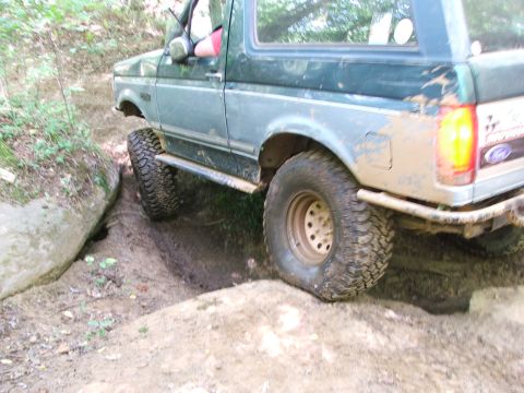 1996 Ford Bronco - Mud-covered Ford Bronco navigates a tight off-road trail with large tires and a green body.