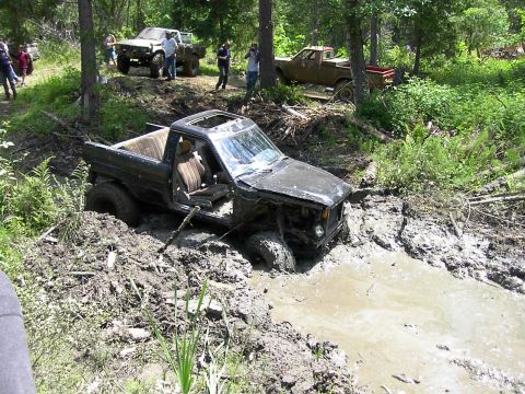 1988 Ford Bronco - Several vehicles and people are gathered in a wooded area with one truck deeply stuck in the mud.
