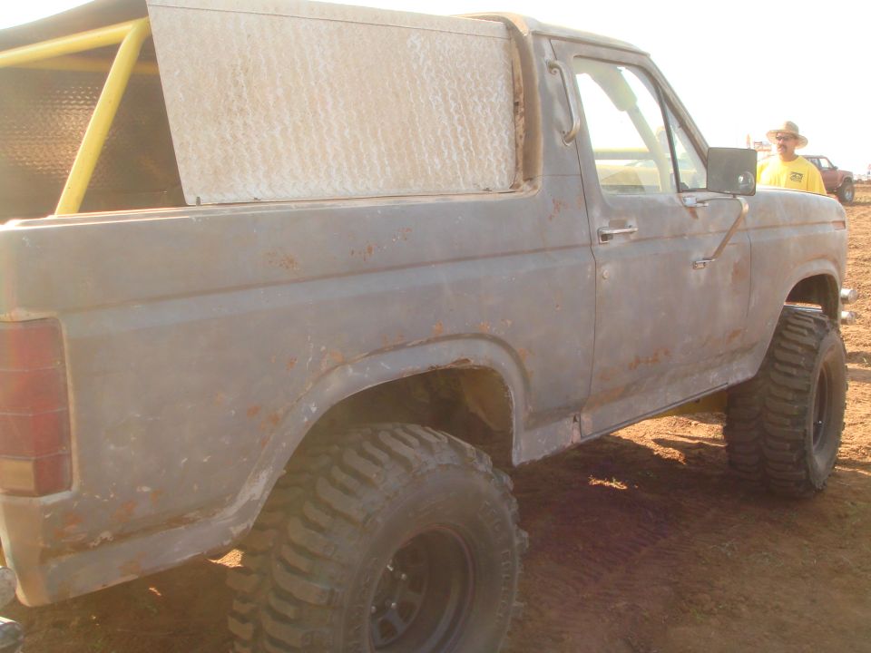 Old Ford Bronco with roll cage and diamond plate rear section, lifted with large tires. Person in hat visible