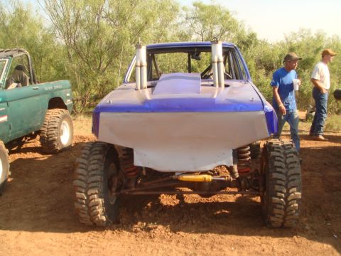 1992 Ford Bronco - A heavily modified off-road vehicle with custom bodywork and exposed suspension sits in a dirt lot next to another off-road vehicle.