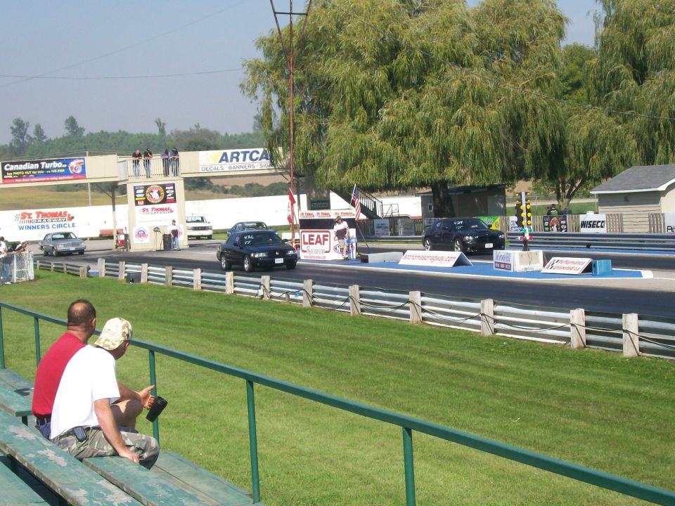 Two dark sedans staged at the starting line of St. Thomas Dragway, seen from grandstands with two spectators.