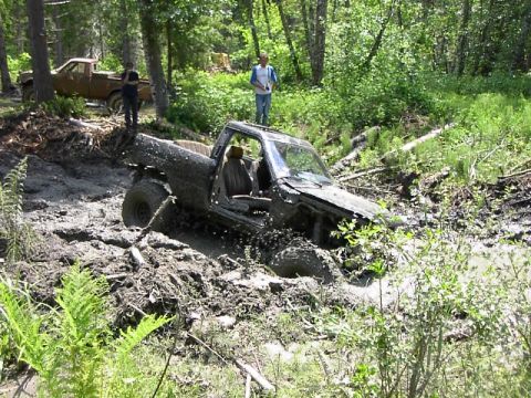 1988 Ford Bronco - This truck is deeply stuck in the mud, with another vehicle visible in the background amidst trees.