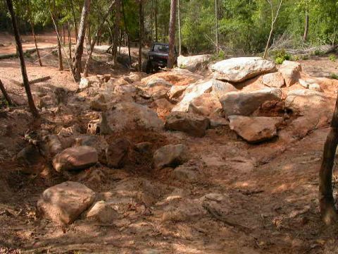 1990 Ford Bronco - A dark off-road vehicle traverses a challenging rocky terrain in a wooded area.
