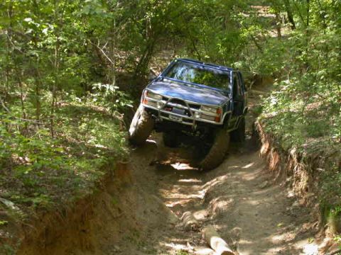 1990 Ford Bronco - A blue 1990 Ford Bronco tackles a challenging off-road course, with its tires gripping the muddy terrain.
