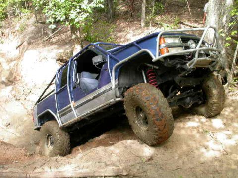 1990 Ford Bronco - A heavily modified blue off-road vehicle navigates a steep, muddy incline.