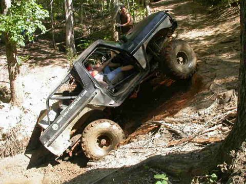 1990 Ford Bronco - A modified off-road vehicle navigates a steep, muddy trail in a wooded area.