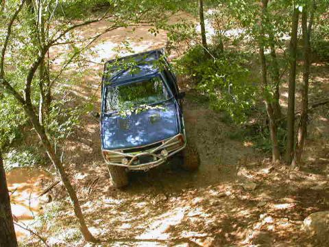 1990 Ford Bronco - A blue 1990 Ford Bronco navigates a wooded trail with a muddy incline.