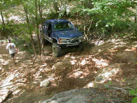 1990 Ford Bronco - A blue off-road truck navigates a steep, rocky incline surrounded by trees.