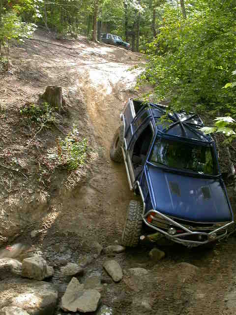 1990 Ford Bronco - Two vehicles navigate a challenging off-road trail through a wooded area.